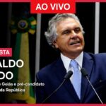 Uma imagem promocional de "Poder Entrevista" para uma entrevista ao vivo com Ronaldo Caiado. Caiado, governador de Goiás e pré-candidato à Presidência da República, está de pé em um púlpito com microfones, com o punho esquerdo fechado em um gesto de ênfase. Ele veste terno azul-escuro, camisa branca e gravata azul-clara estampada. O fundo escuro mostra uma bandeira do Brasil desfocada. O texto na imagem inclui: "PODER ENTREVISTA", "21.jan.2026", "AO VIVO", "ENTREVISTA", "RONALDO CAIADO" e "governador de Goiás e pré-candidato à Presidência da República".