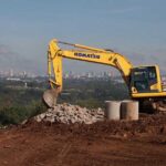 Escavadeira Komatsu amarela operando em área de terraplanagem de aterro sanitário, com pilha de pedras e tubos de concreto em primeiro plano e vista da cidade, realizada pela Prefeitura de Aparecida de Goiânia ao fundo sob céu azul.