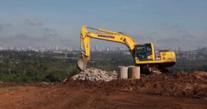 Escavadeira Komatsu amarela operando em área de terraplanagem de aterro sanitário, com pilha de pedras e tubos de concreto em primeiro plano e vista da cidade, realizada pela Prefeitura de Aparecida de Goiânia ao fundo sob céu azul.