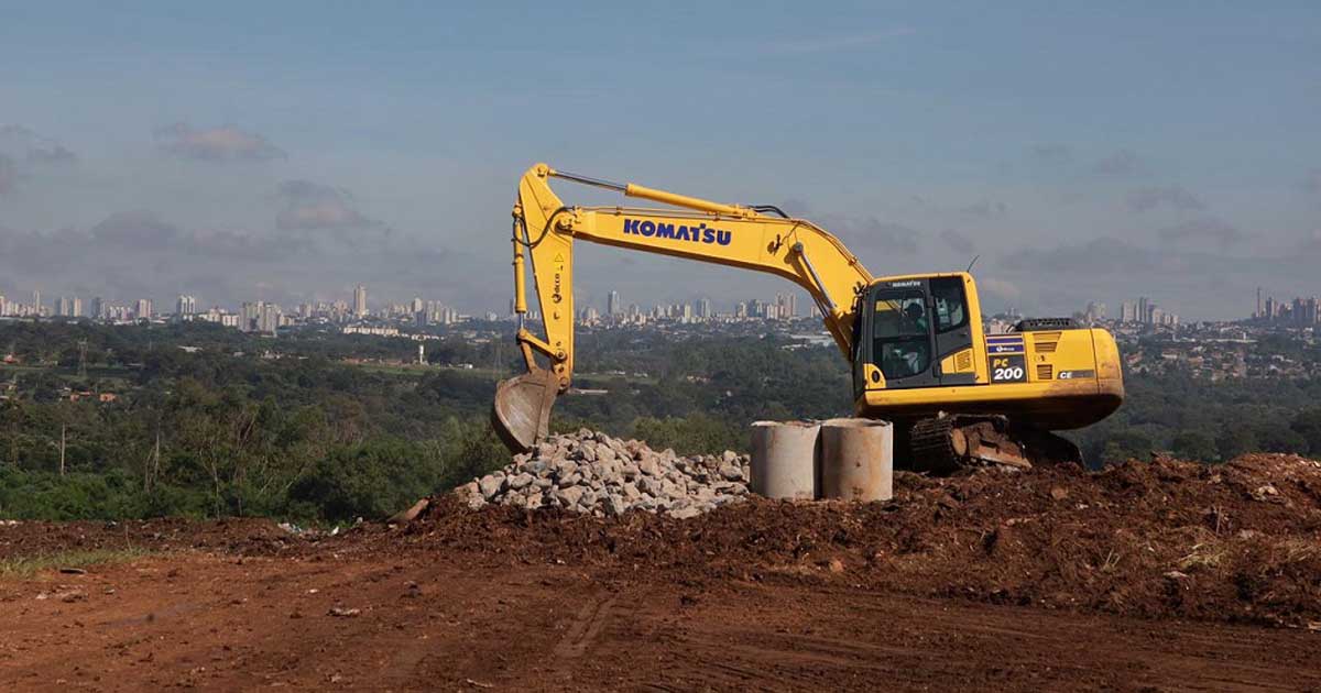 Escavadeira Komatsu amarela operando em área de terraplanagem de aterro sanitário, com pilha de pedras e tubos de concreto em primeiro plano e vista da cidade, realizada pela Prefeitura de Aparecida de Goiânia ao fundo sob céu azul.