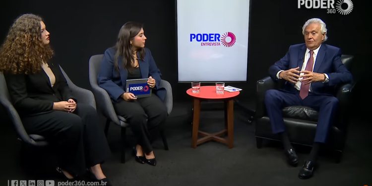 Uma foto de estúdio de uma entrevista do Poder 360. Uma mulher com cabelo encaracolado, Olívia Martins, e terno preto (esquerda) e uma mulher com terno azul-marinho, Juliana Alves, segurando um tablet (centro) ouvem o governador Ronaldo Caiado (direita), um homem mais velho com cabelos brancos e terno azul, falando com as mãos gesticulando. Eles estão sentados em poltronas em torno de uma mesa vermelha com copos de água. Ao fundo, uma tela mostra o logotipo 'PODER ENTREVISTA'. O logotipo 'PODER 360' está no canto superior direito. Barra inferior com URL e ícones de redes sociais.