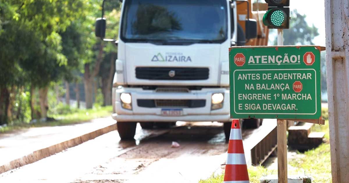 Um caminhão branco com logo da Pedreira Izaíra aproximando-se de uma balança de pesagem rodoviária. À direita, uma placa verde legível com os textos "ATENÇÃO!" e "ANTES DE ADENTRAR NA BALANÇA ENGRENE 1ª MARCHA E SIGA DEVAGAR", junto a cones de trânsito e um semáforo com a luz verde acesa.