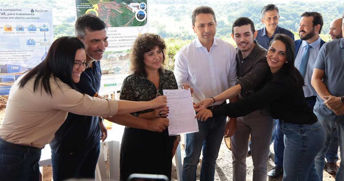 O prefeito de Aparecida de Goiânia, Leandro Vilela, acompanhado de comitiva e técnicos, segura o documento da Licença Ambiental de Operação (LO) em frente a um banner informativo no Aterro Sanitário Municipal.